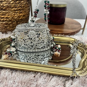 May include: A silver-toned metal clutch with an intricate filigree floral pattern and a hinged lid. The bag features a long silver chain strap adorned with clusters of small red, green, and white beads, along with small shell-like accents.