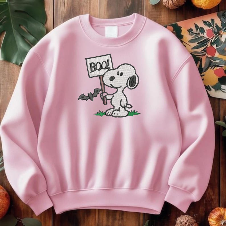 May include: A light pink crewneck sweatshirt featuring an embroidered graphic of the cartoon character Snoopy holding a sign that says BOO with a small bat flying nearby and green grass at the base of the character on the front center.