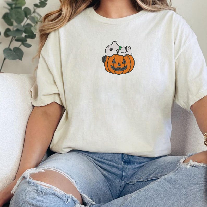 May include: A cream-colored short-sleeve t-shirt featuring a small embroidered graphic on the chest. The design depicts a white cartoon dog with black ears resting on top of an orange jack-o'-lantern pumpkin with a carved face and green stem.