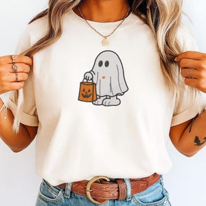 May include: A cream-colored t-shirt featuring an embroidered graphic of a light gray ghost character holding an orange jack-o'-lantern trick-or-treat bucket. The ghost has simple black eyes and a small orange dot on its chest, rendered in a cartoon style.