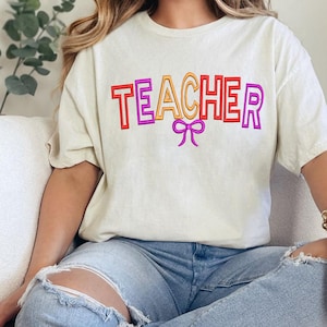Może przedstawiać: Kremowa koszulka z napisem "TEACHER" w kolorowej, obrysowanej czcionce. Słowo jest w kolorze czerwonym, pomarańczowym i fioletowym, z kokardą poniżej. Koszulka ma swobodny styl.