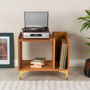 May include: A wooden record player stand with gold-toned metal legs, featuring an open shelf for storage and a vertical side compartment for vinyl records. A record player sits on top, and a stack of books rests on the lower shelf surface.