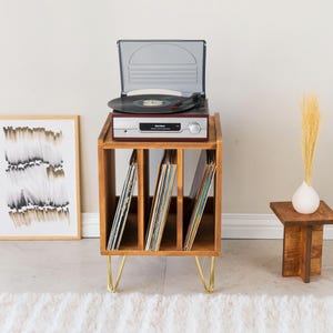 May include: A wooden record player stand with three vertical storage compartments for vinyl records, supported by four gold-toned hairpin legs. A silver and black turntable sits on top, and a small wooden side table holds a white vase with dried grass.