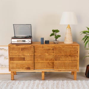 Puede incluir: Un aparador de madera con puertas de listones verticales, coronado con un tocadiscos, dos velas azul oscuro, un pequeño bonsái artificial y una lámpara de mesa con pantalla blanca y base cónica beige con cuello dorado.