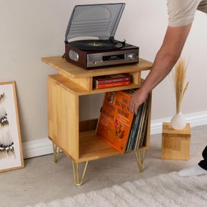 May include: A light wood record player stand with gold-toned hairpin legs, featuring an open storage shelf for vinyl records and a smaller shelf for books. A dark wood turntable with a clear lid sits on top of the stand, which holds several records.