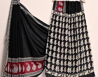 May include: A black saree with a red and white paisley border and a matching black skirt with a white paisley pattern. The saree is draped, and the skirt hangs on a wooden hanger.
