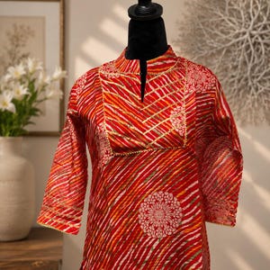 May include: A red tunic with a mandarin collar and three-quarter sleeves. The tunic features a striped pattern in red, white, orange, and green. Gold trim accents the neckline, sleeves, and floral designs.