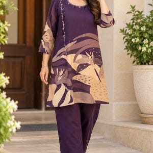 May include: A dark purple tunic and matching trousers set. The tunic features a round neckline with delicate embroidery and a large, abstract floral print in shades of beige, tan, and dark brown across the lower front and sleeves.