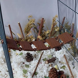 May include: A brown, articulated suspension bridge toy for small animals, featuring a textured wood-grain pattern on each plank. The bridge is suspended between the wire walls of a cage, positioned above white bedding, pinecones, and twigs.