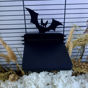 May include: A black platform shelf designed for a wire cage, featuring a silhouette of a bat with pointed ears and outstretched wings attached to the back edge. The shelf has a flat, rectangular surface and is mounted to horizontal wire bars.