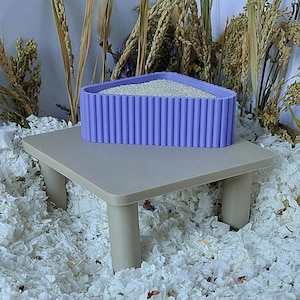 May include: A triangular lavender container with a ribbed exterior texture, filled with fine white granular material, resting on a small, light beige four-legged table. The container features vertical ridges along its entire outer perimeter.