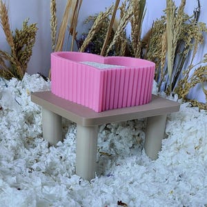 May include: A bright pink heart-shaped container with a ribbed exterior texture, resting on a small, light beige four-legged table. The container features a flat, light-colored interior surface and a distinct heart-shaped silhouette.