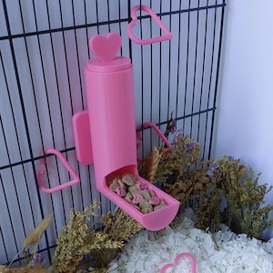May include: A bright pink cylindrical pet food dispenser featuring a heart-shaped handle on the lid. The dispenser is mounted to a black wire cage and contains small, light-colored kibble pieces with pink heart-shaped accents inside the tray.