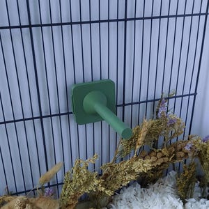 May include: A matte sage green plastic water bottle nozzle attachment featuring a square base plate and a cylindrical spout, designed to be mounted onto a dark metal wire cage grid for small animal hydration and enclosure accessories.