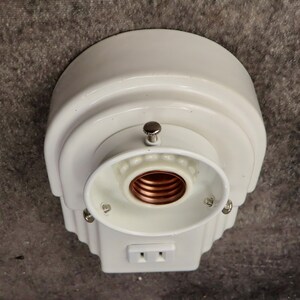 Puede incluir: Una l&aacute;mpara blanca de estilo vintage con un enchufe el&eacute;ctrico integrado. La l&aacute;mpara tiene una parte superior redondeada y un dise&ntilde;o decorativo escalonado. El portal&aacute;mparas es visible y la l&aacute;mpara est&aacute; montada sobre una superficie gris.