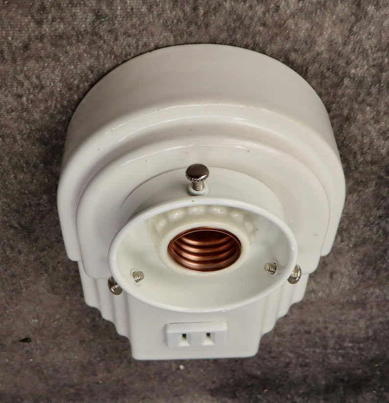 Puede incluir: Una l&aacute;mpara blanca de estilo vintage con un enchufe el&eacute;ctrico integrado. La l&aacute;mpara tiene una parte superior redondeada y un dise&ntilde;o decorativo escalonado. El portal&aacute;mparas es visible y la l&aacute;mpara est&aacute; montada sobre una superficie gris.