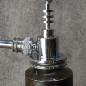 Puede incluir: Primer plano de un objeto met&aacute;lico cromado con una parte superior esf&eacute;rica y anillos apilados. Tiene una base cil&iacute;ndrica y un brazo curvo y texturizado. El art&iacute;culo es probablemente decorativo o funcional, posiblemente un grifo.