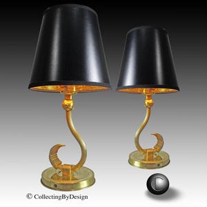 May include: Two elegant table lamps with black shades and gold-colored bases. The lamps feature a curved design with a crescent-shaped detail and a round base. The interior of the shades has a gold finish. The lamps are decorative and stylish.