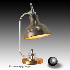 Puede incluir: Una l&aacute;mpara de escritorio de estilo vintage con una pantalla met&aacute;lica y un brazo curvo plateado. La l&aacute;mpara tiene una base redonda de madera con un borde color lat&oacute;n. La pantalla de la l&aacute;mpara es de color met&aacute;lico oscuro y el interior est&aacute; iluminado con una luz c&aacute;lida.