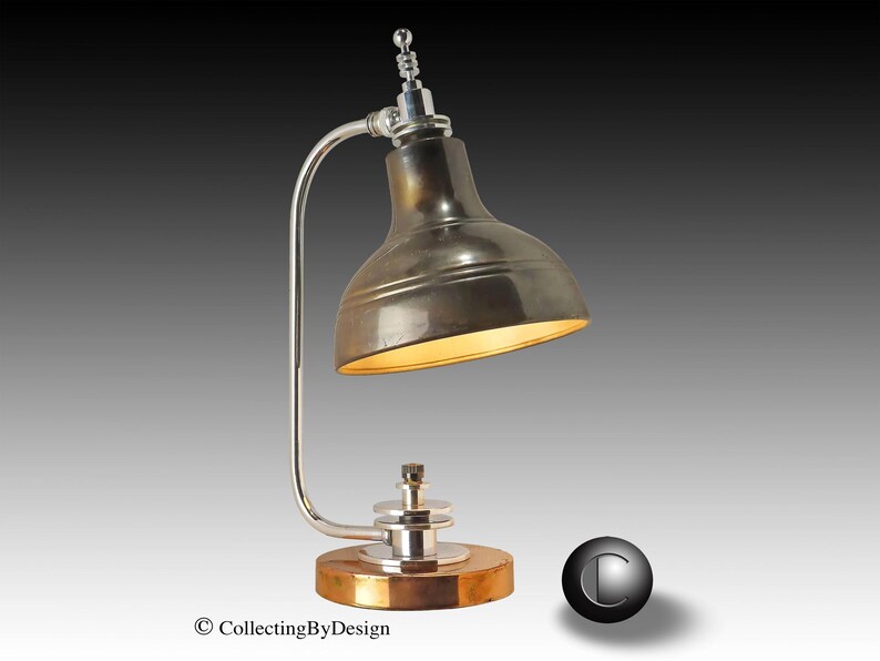 Puede incluir: Una l&aacute;mpara de escritorio de estilo vintage con una pantalla met&aacute;lica y un brazo curvo plateado. La l&aacute;mpara tiene una base redonda de madera con un borde color lat&oacute;n. La pantalla de la l&aacute;mpara es de color met&aacute;lico oscuro y el interior est&aacute; iluminado con una luz c&aacute;lida.