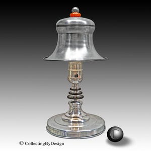 May include: A silver-toned table lamp with a bell-shaped shade and a round base. The lamp features a decorative design with a series of rings and a small orange accent on top. The lamp is designed with a vintage aesthetic.