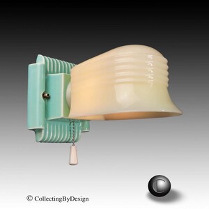 May include: A wall-mounted light fixture featuring a pale mint green rectangular base with vertical ribbed detailing and a cream-colored, semi-translucent glass shade with horizontal ridges. A white pull chain with a small white handle hangs below.