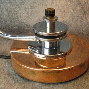 Puede incluir: Primer plano de una l&aacute;mpara de escritorio vintage con base color cobre y brazo cromado. La l&aacute;mpara presenta una base circular, una columna central cromada y un bot&oacute;n negro. El dise&ntilde;o recuerda la &eacute;poca Art D&eacute;co.