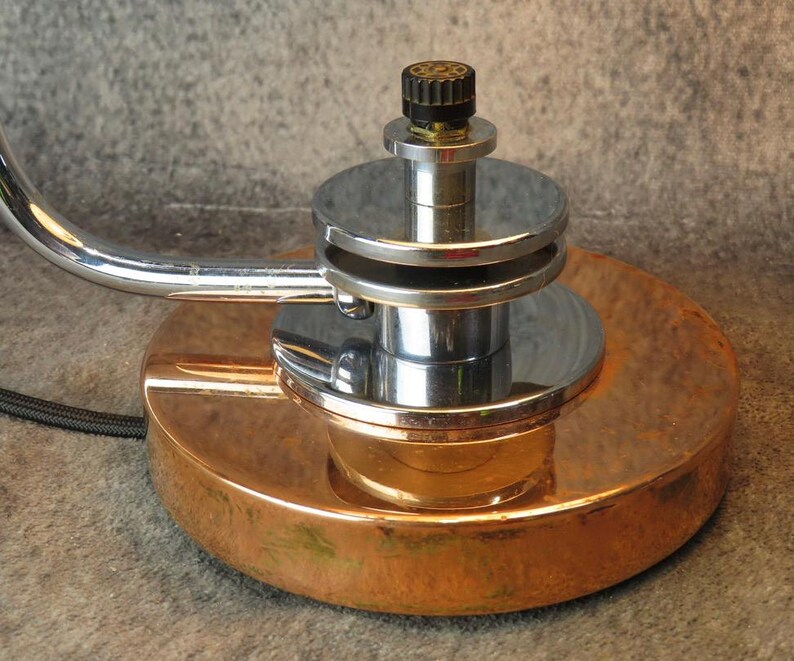 Puede incluir: Primer plano de una l&aacute;mpara de escritorio vintage con base color cobre y brazo cromado. La l&aacute;mpara presenta una base circular, una columna central cromada y un bot&oacute;n negro. El dise&ntilde;o recuerda la &eacute;poca Art D&eacute;co.