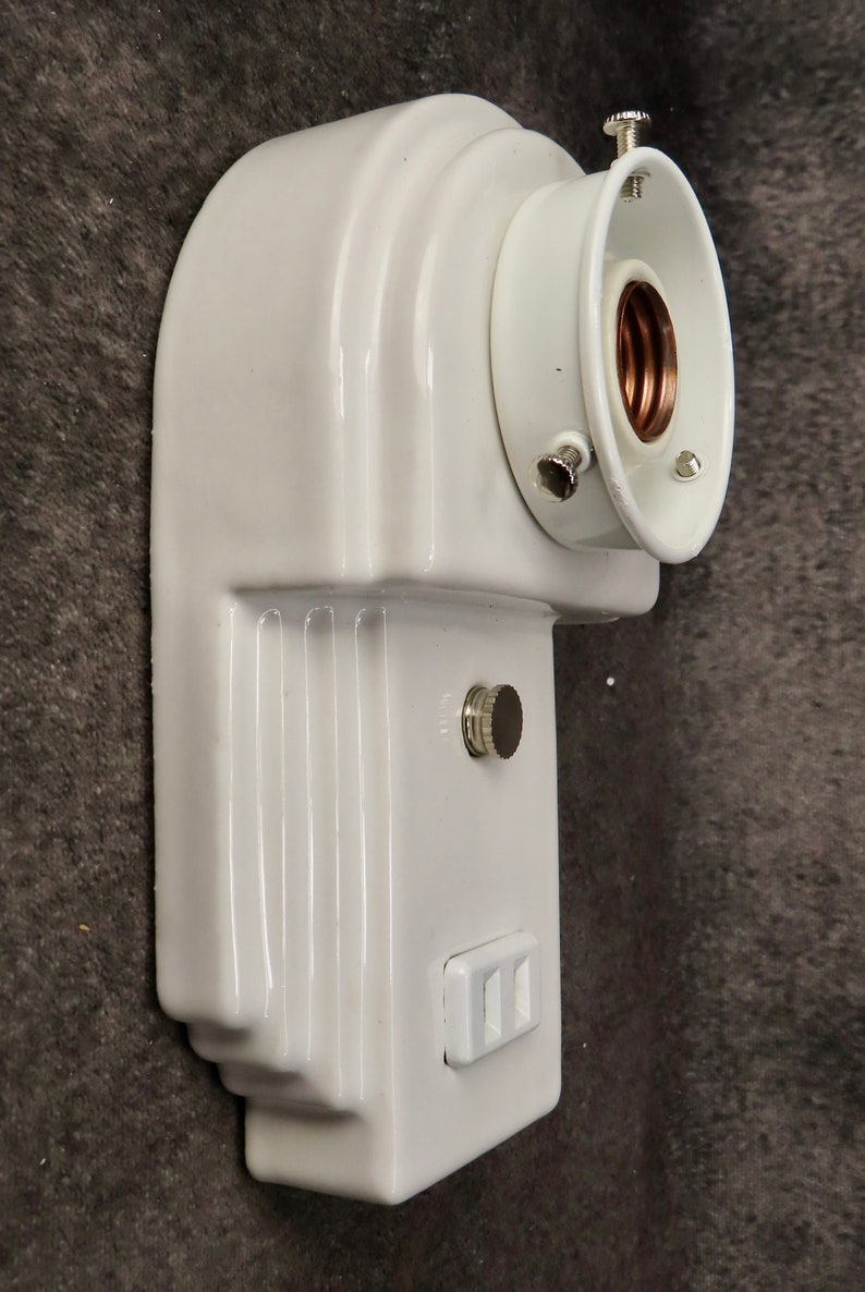 Puede incluir: Una l&aacute;mpara de pared blanca de estilo vintage. La l&aacute;mpara tiene un dise&ntilde;o curvo con un portal&aacute;mparas y un peque&ntilde;o interruptor. La l&aacute;mpara tambi&eacute;n tiene una toma de corriente. La l&aacute;mpara est&aacute; hecha de cer&aacute;mica o porcelana.