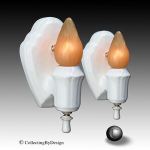 May include: A pair of white ceramic wall sconces featuring a scalloped backplate and a vertical fluted base. Each fixture is topped with a flame-shaped, amber-toned glass bulb that emits a warm glow. The design includes a small white finial.