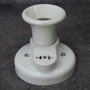 Puede incluir: Un accesorio de luz de cer&aacute;mica blanca con una base circular y una parte superior acampanada. El accesorio tiene una peque&ntilde;a secci&oacute;n rectangular con una toma de dos clavijas. Dos tornillos aseguran el accesorio a una superficie.