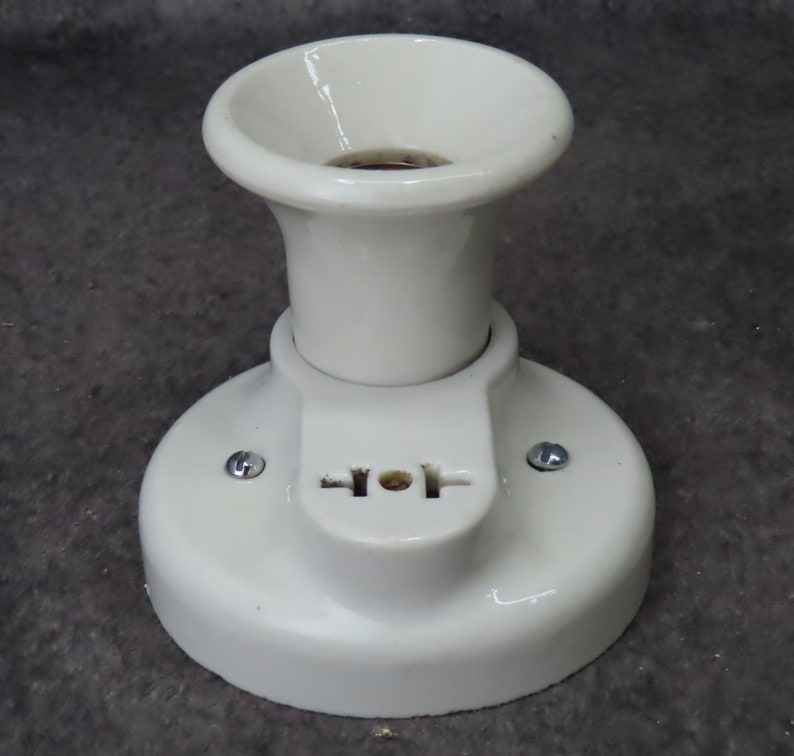 Puede incluir: Un accesorio de luz de cer&aacute;mica blanca con una base circular y una parte superior acampanada. El accesorio tiene una peque&ntilde;a secci&oacute;n rectangular con una toma de dos clavijas. Dos tornillos aseguran el accesorio a una superficie.