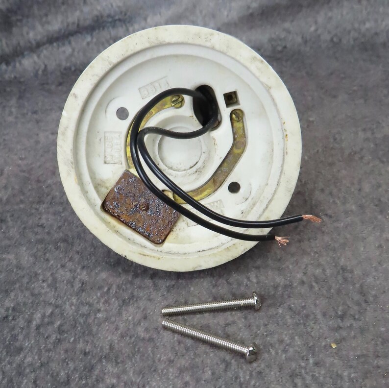 Puede incluir: Un plaf&oacute;n de cer&aacute;mica blanca con cables negros y una placa de metal oxidado. Dos tornillos plateados est&aacute;n colocados debajo. El plaf&oacute;n tiene un dise&ntilde;o circular.