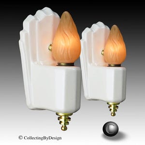 May include: A pair of white ceramic wall sconces featuring stepped, geometric backplates and brass finials at the base. Each fixture holds a single flame-shaped, frosted amber glass bulb that emits a warm, soft glow, creating a focused light.