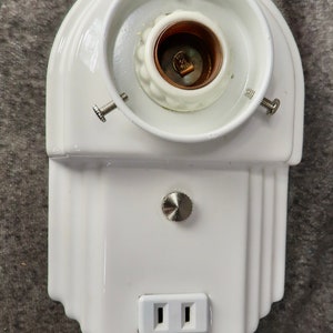 Puede incluir: Una l&aacute;mpara de cer&aacute;mica blanca con un portal&aacute;mparas, un enchufe y un dise&ntilde;o decorativo. La l&aacute;mpara tiene una parte superior redondeada y una base rectangular con un enchufe de dos clavijas.