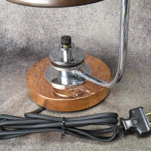 Puede incluir: Una l&aacute;mpara de escritorio vintage con base de cobre y brazo cromado. La l&aacute;mpara tiene un cable de alimentaci&oacute;n y un enchufe negros. El dise&ntilde;o de la l&aacute;mpara es una mezcla de estilos industrial y art d&eacute;co, con enfoque en la funcionalidad.