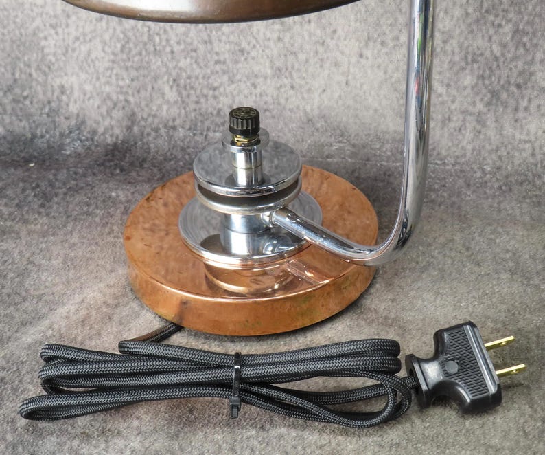 Puede incluir: Una l&aacute;mpara de escritorio vintage con base de cobre y brazo cromado. La l&aacute;mpara tiene un cable de alimentaci&oacute;n y un enchufe negros. El dise&ntilde;o de la l&aacute;mpara es una mezcla de estilos industrial y art d&eacute;co, con enfoque en la funcionalidad.