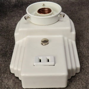 Puede incluir: Una l&aacute;mpara blanca de estilo Art D&eacute;co con un portal&aacute;mparas, un peque&ntilde;o interruptor y una toma de corriente de dos clavijas. La l&aacute;mpara tiene un dise&ntilde;o escalonado con ranuras verticales en los laterales. El portal&aacute;mparas es blanco con interior cobrizo.