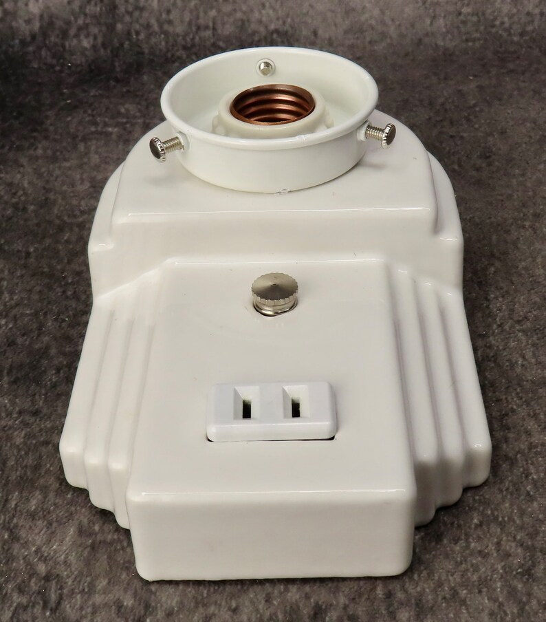 Puede incluir: Una l&aacute;mpara blanca de estilo Art D&eacute;co con un portal&aacute;mparas, un peque&ntilde;o interruptor y una toma de corriente de dos clavijas. La l&aacute;mpara tiene un dise&ntilde;o escalonado con ranuras verticales en los laterales. El portal&aacute;mparas es blanco con interior cobrizo.