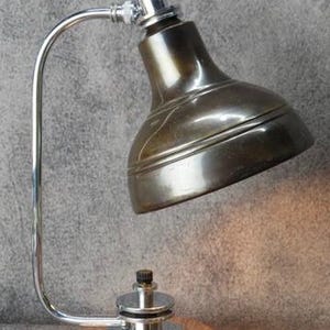 Puede incluir: L&aacute;mpara de escritorio de estilo vintage con una pantalla de bronce met&aacute;lico y un brazo curvo cromado. La l&aacute;mpara tiene una base de color cobre y un cabezal peque&ntilde;o y ajustable. El dise&ntilde;o recuerda la iluminaci&oacute;n industrial.