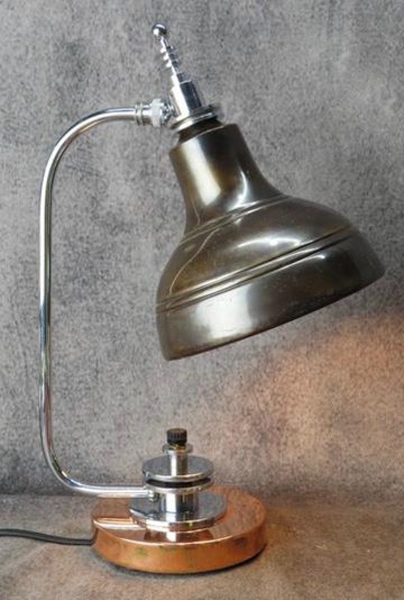 Puede incluir: L&aacute;mpara de escritorio de estilo vintage con una pantalla de bronce met&aacute;lico y un brazo curvo cromado. La l&aacute;mpara tiene una base de color cobre y un cabezal peque&ntilde;o y ajustable. El dise&ntilde;o recuerda la iluminaci&oacute;n industrial.