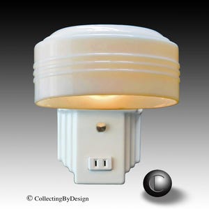 Puede incluir: Una luz de noche blanca vintage con una pantalla redondeada y acanalada y una base cuadrada. La luz de noche tiene una toma de dos clavijas y un peque&ntilde;o interruptor. La luz est&aacute; encendida, emitiendo un brillo c&aacute;lido. Es un art&iacute;culo de colecci&oacute;n.