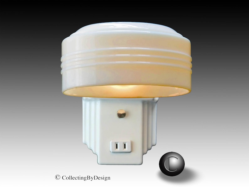 Puede incluir: Una luz de noche blanca vintage con una pantalla redondeada y acanalada y una base cuadrada. La luz de noche tiene una toma de dos clavijas y un peque&ntilde;o interruptor. La luz est&aacute; encendida, emitiendo un brillo c&aacute;lido. Es un art&iacute;culo de colecci&oacute;n.