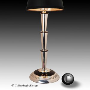 May include: A silver-toned table lamp with a black lampshade and a gold interior. The lamp has a tiered design with a circular base and a slender, elongated body. The lampshade is cylindrical and the base has a black accent.
