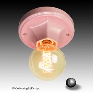 May include: A flush mount ceiling light fixture featuring a glossy, pale pink circular base with two visible mounting screws. A large, clear, spherical Edison-style light bulb with a visible glowing filament is attached to the center of the base.