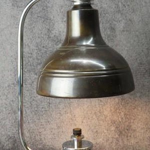 Puede incluir: Una l&aacute;mpara de escritorio de estilo vintage con una pantalla de bronce met&aacute;lico y un brazo cromado curvado. La l&aacute;mpara presenta una base circular de cobre y un mecanismo ajustable plateado. El dise&ntilde;o recuerda a la iluminaci&oacute;n industrial.