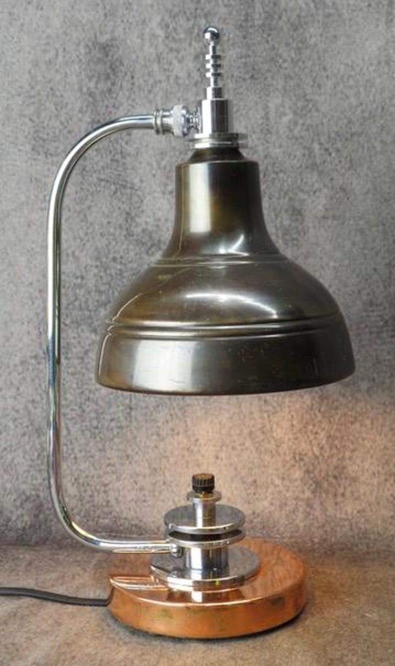 Puede incluir: Una l&aacute;mpara de escritorio de estilo vintage con una pantalla de bronce met&aacute;lico y un brazo cromado curvado. La l&aacute;mpara presenta una base circular de cobre y un mecanismo ajustable plateado. El dise&ntilde;o recuerda a la iluminaci&oacute;n industrial.