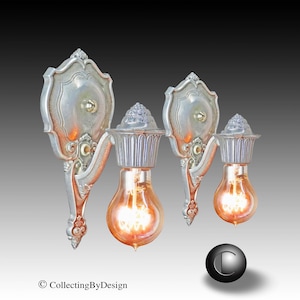 May include: Two ornate silver wall sconces with exposed Edison-style light bulbs. The sconces have a decorative, vintage design with a shield-shaped backplate and intricate detailing. The bulbs emit a warm, orange glow.