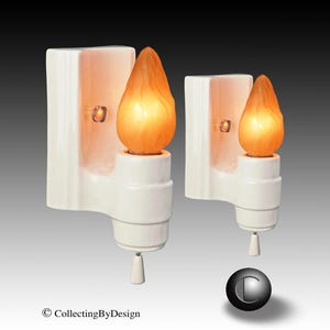 May include: Two white wall sconces with a flame-shaped light bulb. The sconces have a rectangular backplate and a decorative base. The light bulbs are orange and emit a warm glow. The sconces are designed for indoor use.