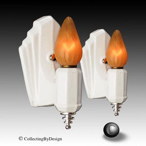 May include: A pair of white ceramic wall sconces featuring a geometric, stepped backplate design. Each fixture holds a single flame-shaped, frosted amber glass bulb that emits a warm glow. The base of each light includes a small, silver-toned metal finial.